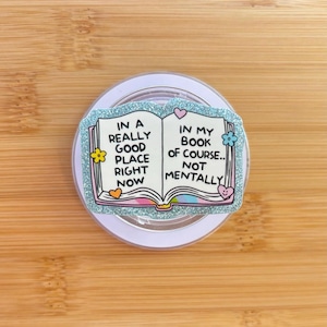 May include: A glittery light blue phone grip with the text "IN A REALLY GOOD PLACE RIGHT NOW" on the left page and "IN MY BOOK OF COURSE.. NOT MENTALLY" on the right page. The design includes flowers and hearts.