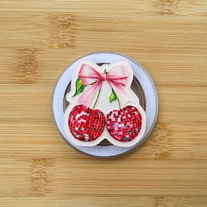 May include: A round, white and clear phone grip with a cherry design. The cherries are red with a mosaic pattern, and the design includes a pink bow and green leaves. The grip is on a wooden surface.