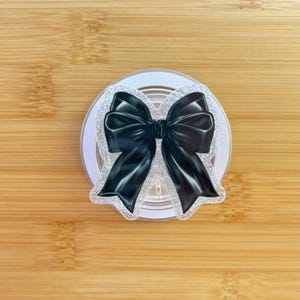 Puede incluir: Un accesorio negro en forma de lazo con un borde de purpurina plateada. El lazo está centrado en una base circular blanca. El fondo es una superficie de madera marrón claro.