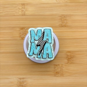 Può includere: Un supporto per telefono pop-up azzurro con la scritta "MAMA" in grassetto e contornata. Un fulmine a scacchi in bianco e nero è centrato tra le due parole. L'impugnatura è su una base circolare bianca, su uno sfondo di legno.