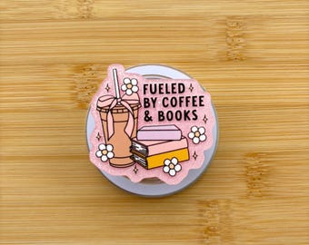 May include: A pink and white pop socket with the text "Fueled by Coffee & Books." The design includes a coffee cup, books, and floral accents. The pop socket is on a light brown wooden surface.