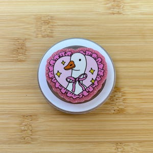 May include: A circular phone grip featuring a white goose illustration centered within a pink heart frame. The goose has an orange beak and a pink bow around its neck. The heart is surrounded by a ruffled pink border with a glittery finish and yellow star accents.