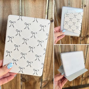 May include: A white e-reader cover with a black bow pattern. The cover has a grid design with small black bows. The image also shows a clear case with a bow pattern and a stand for the e-reader.