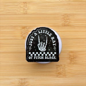 May include: A black and white sticker featuring the text "JUST A LITTLE RAY OF PITCH BLACK" and a skeleton hand in a rock and roll pose. The sticker is on a white circular base, set against a wooden surface.