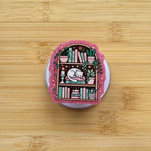 May include: A glittery sticker featuring a dark brown bookshelf illustration with three shelves. The shelves contain various books, potted plants, and a white cat sleeping on a stack of books. The sticker has a pink, sparkling border.