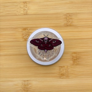 Pode incluir: Um estojo compacto redondo e branco com um design decorativo de mariposa. A mariposa é bordô com detalhes de caveira e olhos. O estojo compacto tem uma parte superior transparente com glitter e um design de corrente prateada.