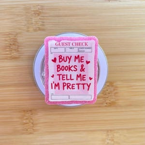 May include: A pink glittery guest check with the text "BUY ME BOOKS & TELL ME I'M PRETTY" in red. The guest check has a white border and is on a white circular base.