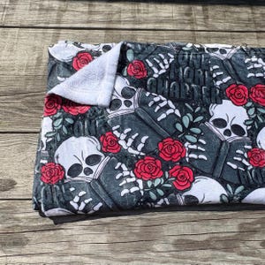 May include: A folded, dark gray blanket with a repeating pattern of white skulls, skeleton hands, and red roses. The text "ONE MORE QUARTER" is visible. The blanket has a white, folded edge.