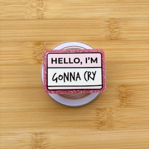 以下が含まれることがあります： 「HELLO, I'M GONNA CRY」の黒い文字が入ったピンクのグリッターフォングリップ。フォングリップは白い背景とピンクのグリッターボーダーが付いています。背景は明るい茶色の木製の表面です。