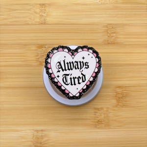 May include: A white heart-shaped pop socket with the words "Always Tired" in black gothic script. The heart has a black and pink scalloped border and is set on a white base.