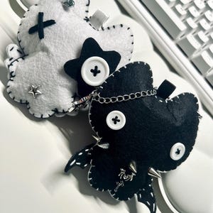 Puede incluir: Dos broches de fieltro hechos a mano. Uno es blanco con una cruz negra, una estrella y un imperdible. El otro es negro con una estrella, una cadena, botones y púas plateadas. Ambos tienen costuras blancas.
