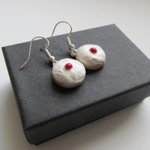 May include: Donut-shaped earrings with white icing and a red dot, presented on a black box. The earrings have silver-coloured hooks. The miniature donuts are a fun, quirky accessory, perfect for adding a touch of whimsy.