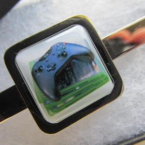 May include: A gold-coloured tie clip with a square emblem. The emblem features a blue video game controller design on a green and white background, framed by a black square.
