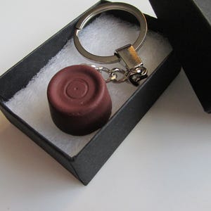 May include: A dark brown keychain charm with a circular design, attached to a silver keyring. The charm is presented in a small black gift box with white padding.