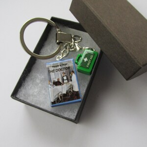 May include: A silver keyring with three charms: a miniature book titled "THE DOCTOR", a green first aid kit, and a silver ring. The keyring is in a black box with a brown lid.