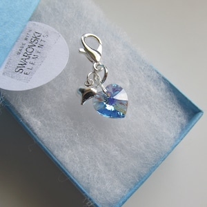 May include: A silver charm featuring a light blue heart-shaped crystal and a small silver heart charm. The charm is made with Swarovski elements and comes in a blue box with white tissue paper. "Made with Swarovski Elements" is printed on a white label.