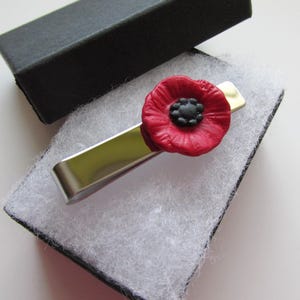 May include: A silver tie bar with a red poppy flower charm. The poppy has a black centre.