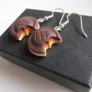 May include: A pair of earrings shaped like chocolate chip cookies with a bite taken out of them. The earrings are silver and have a dark brown and light brown colour scheme.