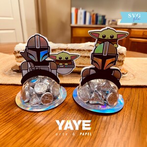 Puede incluir: Dos portadores de caramelos con temática de Mandalorian con chocolates envueltos en papel de aluminio plateado. Cada portador presenta un recorte del Mandaloriano y Grogu, con una cúpula transparente. La base es iridiscente. El texto "YAYE arte & PAPEL" está en la parte inferior.