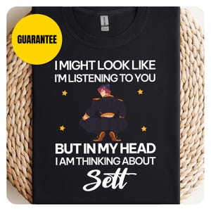 May include: Black t-shirt with the text "I MIGHT LOOK LIKE I'M LISTENING TO YOU BUT IN MY HEAD I AM THINKING ABOUT Sett" and a cartoon illustration. A yellow "GUARANTEE" circle is in the upper left corner.