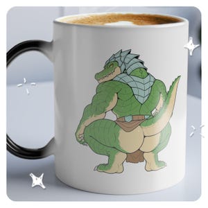 Op de afbeelding: Witte keramische mok met een groene antropomorfe alligator in een hurkende houding. Het personage draagt een zilveren helm en harnas. De mok heeft een zwart handvat en is gevuld met een bruine vloeistof.