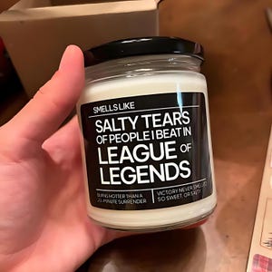 May include: A white candle with a black label that reads "Smells Like Salty Tears of People I Beat in League of Legends". The label also says "Burns hotter than a 20-minute surrender" and "Victory never smelled so sweet, or salty".