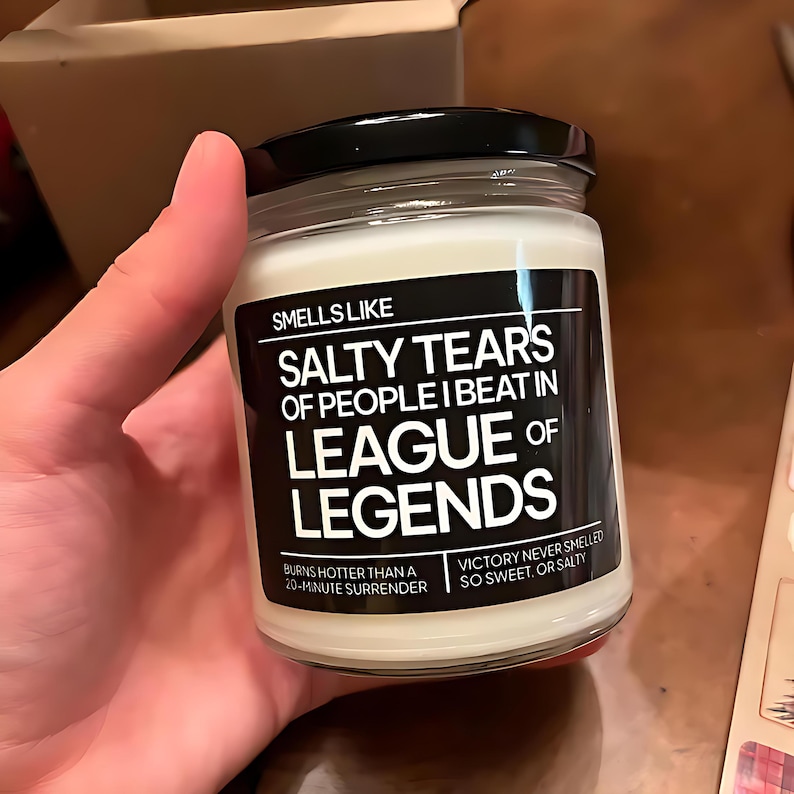 May include: A white candle with a black label that reads "Smells Like Salty Tears of People I Beat in League of Legends". The label also says "Burns hotter than a 20-minute surrender" and "Victory never smelled so sweet, or salty".