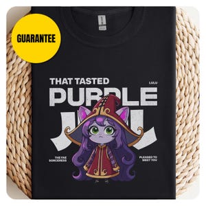 Könnte beinhalten: Schwarzes T-Shirt mit einer weißen Grafik eines Comic-Charakters mit lila Haaren und einem roten Hut. Der Text "THAT TASTED PURPLE" befindet sich über dem Charakter und "LULU THE FAE SORCERESS" unter dem Charakter.