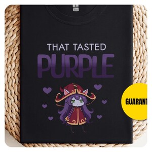 May include: Black t-shirt with the text "THAT TASTED PURPLE" in white and purple. A cartoon character with purple hair and a red hat is featured below the text, surrounded by purple hearts.