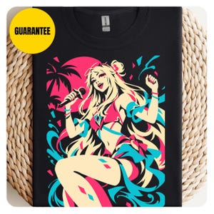 May include: A black t-shirt featuring a vibrant graphic print of a blonde character holding a microphone. The design uses a color palette of hot pink, cyan, and cream, depicting a tropical scene with palm trees and a large sun. The word GUARANTEE is printed in black on a yellow circle in the top left corner.