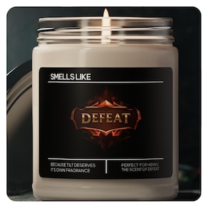Puede incluir: Una vela encendida en un tarro de cristal con una etiqueta negra. La etiqueta dice "SMELLS LIKE DEFEAT" en letras doradas. Debajo del texto están las frases "BECAUSE TILT DESERVES IT'S OWN FRAGRANCE" y "PERFECT FOR HIDING THE SCENT OF DEFEAT."