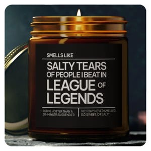 May include: A brown glass candle jar with a gold lid and a black label that reads "Smells Like Salty Tears of People I Beat in League of Legends". The label also includes the text "Burns hotter than a 20-minute surrender" and "Victory never smelled so sweet, or salty".