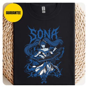 May include: Black t-shirt featuring a blue graphic of a character playing a stringed instrument. The word "SONA" is displayed in stylized blue lettering above the character. The character has long blue hair and is wearing a blue and white outfit.