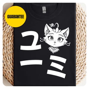 May include: Black t-shirt with a white graphic of a cat's face and Japanese characters. A yellow circle with the word "GUARANTEE" is in the upper left corner. The shirt is displayed on a woven surface.