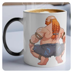 Può includere: Una tazza da caffè in ceramica bianca con un manico nero. La tazza presenta un'illustrazione di cartone animato di un uomo muscoloso con i capelli rossi e la barba, che indossa un perizoma blu e nero, accovacciato con le spalle al pubblico.