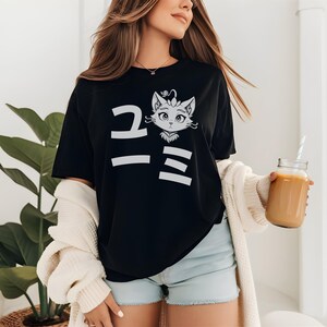 May include: Black t-shirt with a white graphic of a cat face and Japanese characters. The cat has large eyes and a crown. The shirt is a casual style.