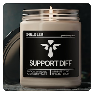 Könnte beinhalten: Eine braune Sojawachskerze mit einem schwarzen Etikett, auf dem "Smells Like Support Diff" steht und ein weißer Engel mit einem Krawattenlogo abgebildet ist. Das Etikett trägt außerdem die Aufschrift "Für diejenigen, die mehr schützen als farmen" und "Eine Hommage an die unbesungenen Helden".