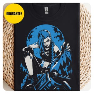 Puede incluir: Una camiseta negra con una ilustración gráfica de un personaje con cabello azul y trenzas, vistiendo un top y pantalones negros, sentado sobre un objeto mecánico con forma de tiburón frente a una gran luna azul. El texto GUARANTEE aparece en un círculo amarillo.