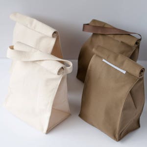 Puede incluir: Tres bolsas de almuerzo, dos son de lona blanca y una de lona marr&oacute;n. La bolsa marr&oacute;n tiene una correa blanca.
