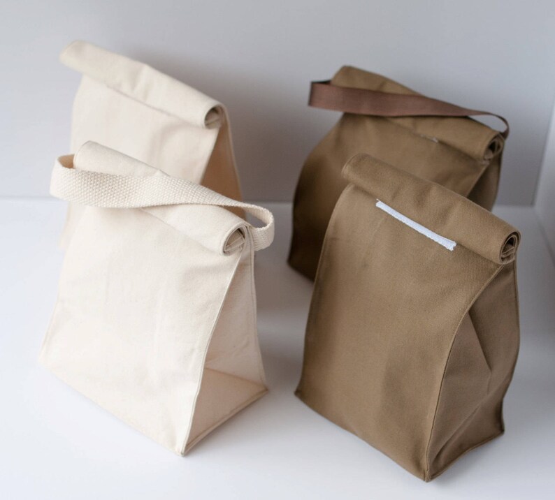 Puede incluir: Tres bolsas de almuerzo, dos son de lona blanca y una de lona marr&oacute;n. La bolsa marr&oacute;n tiene una correa blanca.