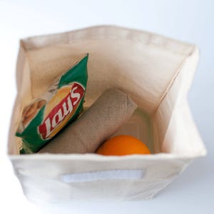 Puede incluir: Una bolsa de almuerzo de lona blanca con cierre de cremallera. La bolsa est&aacute; llena de una bolsa de papas fritas Lay's, un pa&ntilde;o enrollado y una naranja.