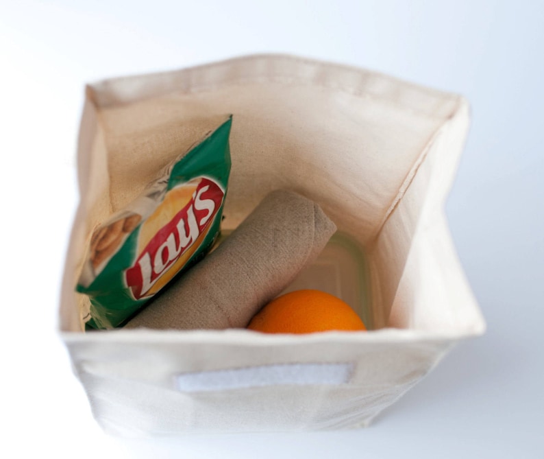 Puede incluir: Una bolsa de almuerzo de lona blanca con cierre de cremallera. La bolsa est&aacute; llena de una bolsa de papas fritas Lay's, un pa&ntilde;o enrollado y una naranja.