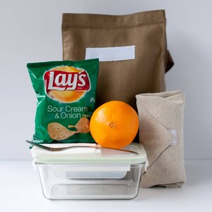 Puede incluir: Una bolsa de almuerzo de lona marr&oacute;n con una etiqueta blanca, una bolsa de papas fritas Lay's Sour Cream & Onion, una naranja, un recipiente de vidrio con tapa y una servilleta de tela beige.