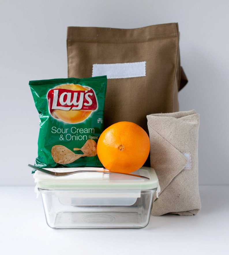 Puede incluir: Una bolsa de almuerzo de lona marr&oacute;n con una etiqueta blanca, una bolsa de papas fritas Lay's Sour Cream & Onion, una naranja, un recipiente de vidrio con tapa y una servilleta de tela beige.