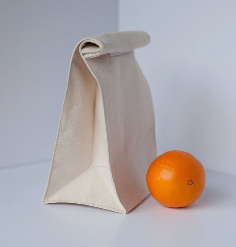 Puede incluir: Una bolsa de almuerzo de lona blanca con la parte superior doblada. La bolsa est&aacute; sobre una superficie blanca junto a una naranja.