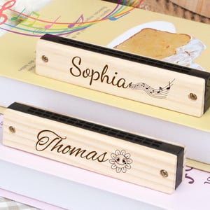 Puede incluir: Dos armónicas de madera con nombres personalizados, "Sophia" y "Thomas". Las armónicas están decoradas con notas musicales y un sol sonriente.
