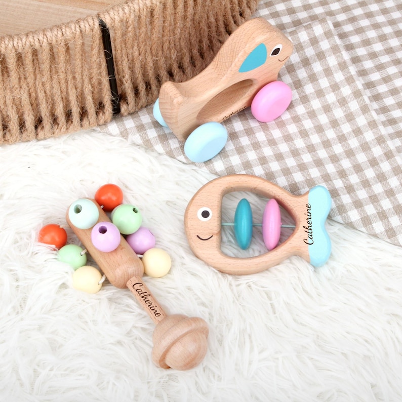 May include: Wooden baby toys including a plane, a fish-shaped rattle, and a rattle with colorful beads. The plane has blue and pink accents. The fish rattle has blue and pink beads. The name "Catherine" is printed on the toys.