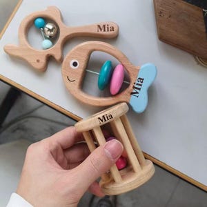 May include: Wooden baby toys, including a fish-shaped rattle with blue and pink beads, a fish-shaped rattle with a blue tail, and a cage-shaped rattle. Each toy is engraved with the name "Mia".
