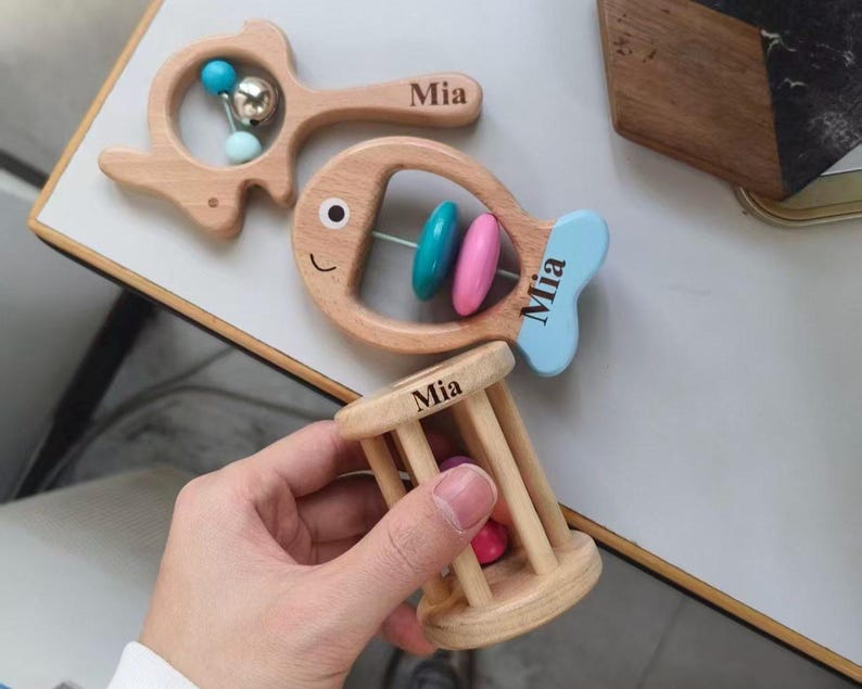 May include: Wooden baby toys, including a fish-shaped rattle with blue and pink beads, a fish-shaped rattle with a blue tail, and a cage-shaped rattle. Each toy is engraved with the name "Mia".