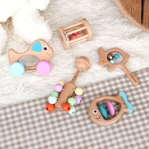 May include: A collection of wooden baby toys, including a fish-shaped rattle, a car-shaped toy, and a rolling cylinder. The toys are made of natural wood and feature pastel-colored accents. Each toy has the name "Catherine" engraved on it.